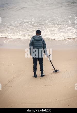 Un seul homme non identifiable se tient debout sur une plage de sable, tenant un détecteur de métaux alors qu'il balaye le rivage à la recherche de trésors cachés Banque D'Images