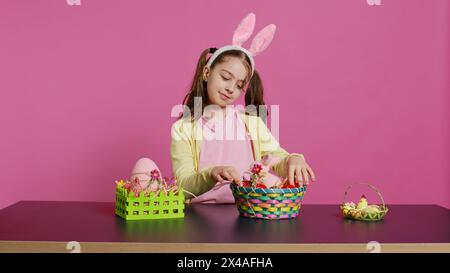 Jeune fille excitée arrangeant des œufs peints dans un panier pour préparer la célébration des vacances de pâques, créant des arrangements festifs. Enfant heureux ludique avec des oreilles de lapin, activité créative. Caméra B. Banque D'Images