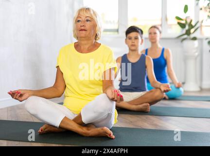 Vieille femme pratiquant la pose demi-lotus du yoga dans la salle de fitness légère Banque D'Images