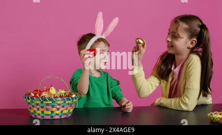 Enfants doux frappant des œufs ensemble pour la tradition de pâques en studio, jouant un jeu de vacances saisonnier sur fond rose. Adorables enfants s'amusant avec des décorations festives. Caméra B. Banque D'Images