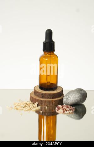 Une bouteille brune d'une huile essentielle sur une surface spéculaire assise sur le bois et entourée de rochers, coquillages et avoine. Banque D'Images