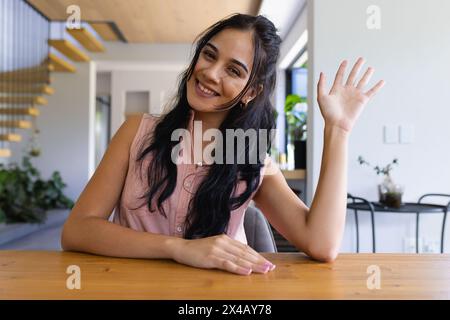 Jeune femme biraciale assise à table, agitant et souriant à la maison sur un appel vidéo Banque D'Images