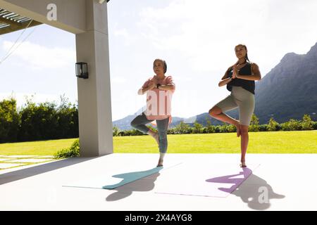 Grand-mère asiatique et petite-fille adolescente biraciale pratiquant le yoga à l'extérieur. Les deux debout sur des tapis, se concentrant sur des poses dans un environnement serein, inaltérer Banque D'Images