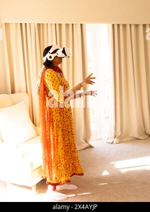 Jeune femme indienne portant un casque de réalité virtuelle à la maison, debout près de la fenêtre. Portant une robe florale jaune et des pantoufles blanches, explorant le monde virtuel Banque D'Images