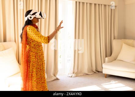 Jeune femme indienne portant un casque de réalité virtuelle à la maison, debout dans une pièce lumineuse. Porter une tenue traditionnelle jaune avec des motifs rouges, profiter de moi Banque D'Images