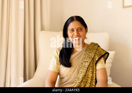 Une femme indienne d'âge moyen en tenue traditionnelle, souriante à la maison. Portant des bijoux en or, elle a de longs cheveux noirs et un teint clair, inaltérés Banque D'Images