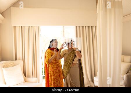 Mère indienne et fille adolescente essayant un casque de réalité virtuelle à la maison. Les deux sportifs cheveux noirs, jeune femme en robe jaune vif, la maturité Banque D'Images