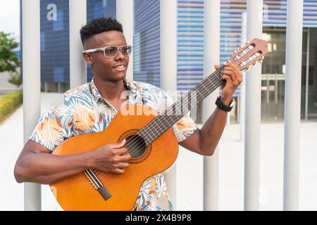 Jeune homme dans des lunettes joue de la guitare acoustique, portant une chemise tropicale dans un cadre urbain. Banque D'Images