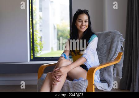Jeune femme biraciale assise dans une chaise, souriante à la maison, espace de copie. Elle a de longs cheveux foncés, porte des vêtements décontractés et aucun maquillage visible, inchangé Banque D'Images