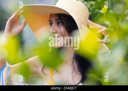 Jeune femme biraciale portant un chapeau à large bord, souriant dehors parmi les feuilles vertes. Elle, ayant les cheveux foncés et la peau brun clair, portant un haut jaune, en Banque D'Images