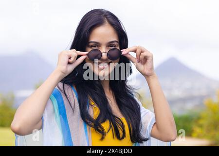 Jeune femme biraciale tenant des lunettes de soleil, souriant dehors à la maison. Elle a de longs cheveux foncés, portant un haut jaune et un châle rayé, inaltérés Banque D'Images