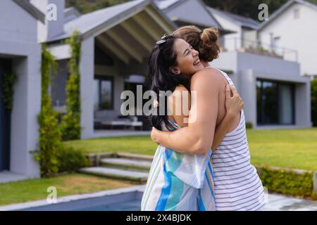 Couple diversifié embrassant à l'extérieur près de la piscine, maison sur fond bleu. Elle, ayant les cheveux foncés, le tenant étroitement, lui dans une chemise rayée, le regardant Banque D'Images