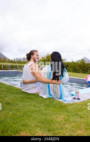 Couple diversifié appréciant la compagnie de l'autre à l'extérieur par piscine, espace de copie. Elle, avec de longs cheveux noirs, et lui, la barbe sportive et les cheveux bouclés, sont détendus Banque D'Images