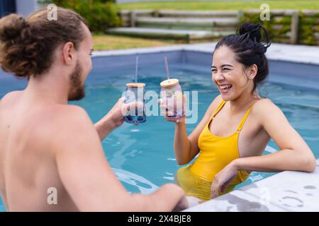 Couple diversifié profitant de boissons à l'extérieur dans la piscine, riant ensemble. Elle, avec les cheveux foncés dans un chignon, et lui, les cheveux bouclés, partagent un Mo joyeux Banque D'Images