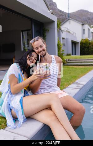 Femme biraciale et homme caucasien, couple, à l'extérieur, assis près de la piscine, tenant des verres. Les deux vêtements d'été décontractés sportifs, elle avec les cheveux noirs, lui avec Banque D'Images