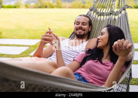 Couple diversifié se relaxant à l'extérieur dans un hamac, homme tenant un smartphone. Les deux avec les cheveux foncés, elle avec la peau brun clair et lui avec la peau claire, appréciant qu Banque D'Images