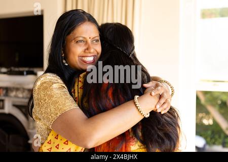 Mère indienne en top or, embrassant sa fille adolescente à la maison, les deux souriant brillamment. Longs cheveux noirs encadrant son visage, rouge à lèvres rehaussant son joyfu Banque D'Images
