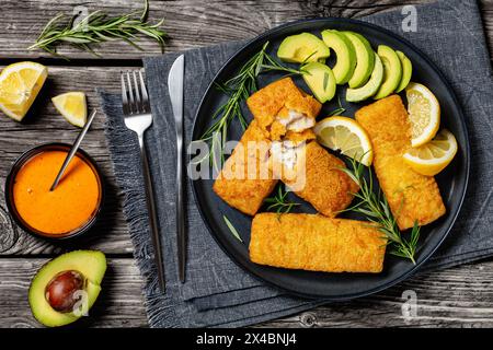 filet de pollock d'alaska croustillant cuit au four sur plat noir avec des tranches de romarin, de citron et d'avocat sur une table en bois rustique avec des couverts et de la crème cajun Banque D'Images