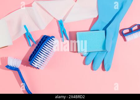 Produit en plastique de nettoyage de maison sur fond rose. Gant en caoutchouc bleu, différents types de brosse, rouleau de papier et pinces à linge décorés. Vue de dessus, scène Banque D'Images