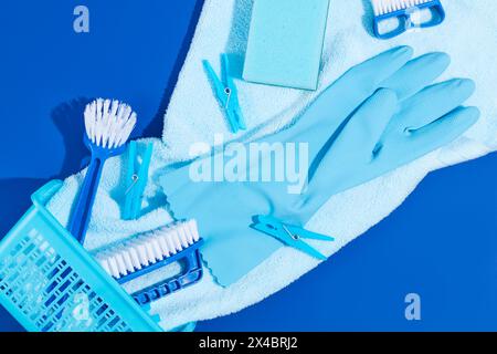 Un panier en plastique avec des fournitures de nettoyage décoré sur fond bleu. Brosses, gants en caoutchouc et pinces à linge affichés. Scène de nettoyage publicitaire Banque D'Images