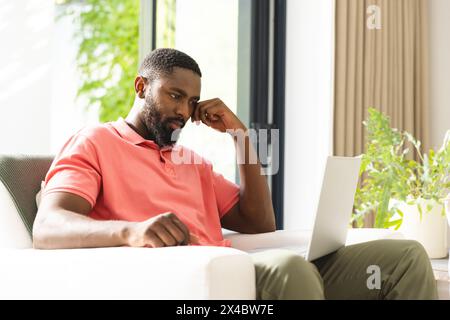 À la maison, homme afro-américain en chemise rose et pantalon vert examinant l'écran de l'ordinateur portable. Avec les cheveux noirs courts, il est assis confortablement, entouré d'un confortable Banque D'Images