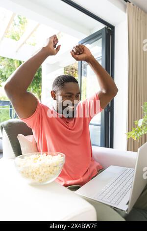 A la maison, homme afro-américain portant une chemise rose, célébrant devant un ordinateur portable. Tenant un bol de pop-corn, arborant une barbe et des cheveux bouclés courts, Banque D'Images