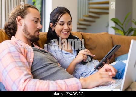 Couple diversifié assis à la maison, regardant un ordinateur portable ensemble. Femme biraciale tenant un smartphone, homme caucasien tapant, les deux portant des vêtements décontractés, unal Banque D'Images