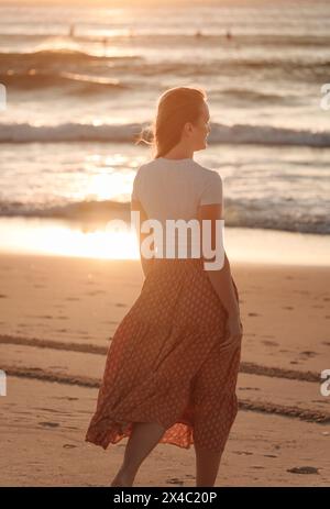 Une femme belle, solitaire et réfléchie au bord de la mer, debout regardant l'horizon, sur la plage à l'aube rougeâtre du lever du soleil, avec son flowin Banque D'Images