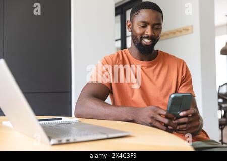 Homme afro-américain en chemise orange, barbu, sourit à son téléphone à la maison. Utilisant un smartphone, avec un ordinateur portable à proximité, il respire une ambiance détendue, unalte Banque D'Images