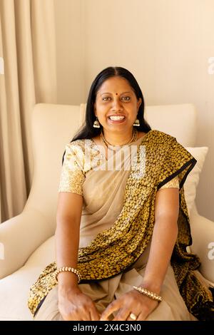 Femme indienne mature portant du sari doré et noir assis, souriant à la maison. Elle, ayant les cheveux foncés et les yeux bruns, portant des bijoux, profitant du moment calme Banque D'Images