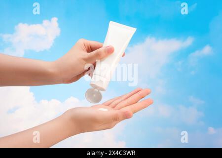 Contre le ciel bleu avec blanc nuageux, les mains de la femme appliquent la crème de tube cosmétique blanc sans étiquette. Scène minimale pour la publicité cosmet Banque D'Images