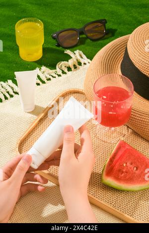 Sur le tapis de pique-nique avec la tasse d'eau fraîche, chapeau, lunettes de soleil affiché bouteille cosmétique blanche. Les mains de la femme se préparent à ouvrir un tube cosmétique et à l'utiliser. M Banque D'Images