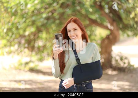 Belle femme rousse avec un bras dans une écharpe sourit tout en tenant son smartphone Banque D'Images