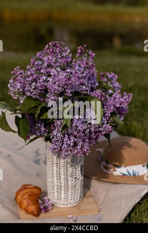 Bouquet de lilas dans un vase en osier, croissant et chapeau. Pique-nique de printemps. Photo de haute qualité Banque D'Images