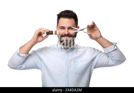 Beau jeune homme avec moustache tenant des ciseaux et une brosse à raser sur fond blanc Banque D'Images