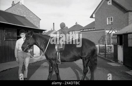 Années 1960, historique, à l'extérieur dans une école d'équitation de banlieue, un enfant en bas âge assis sur un cheval près des écuries, la tête du cheval étant tenue par une adolescente, Angleterre, Royaume-Uni. Banque D'Images