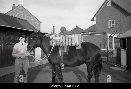 Années 1960, historique, à l'extérieur dans une école d'équitation de banlieue, un enfant en bas âge assis sur un cheval près des écuries, la tête du cheval étant tenue par une adolescente, Angleterre, Royaume-Uni. Banque D'Images