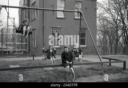 années 1960, historique, écolières devant un bâtiment scolaire dans une aire de jeux, deux assis sur les sièges de la balançoire traditionnelle à mailles à chaîne et une de la fille chevauchant haut dans les airs. Banque D'Images