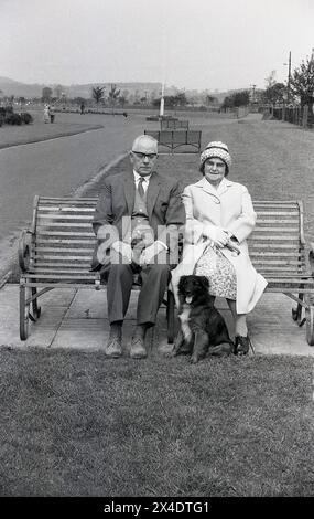 années 1960, historique, un couple âgé, formellement habillé, le gentleman en costume et cravate, la dame en manteau et chapeau, assis ensemble sur un banc de parc en métal avec leur chien après l'avoir emmené pour une promenade i. Banque D'Images