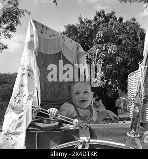 années 1960, historique, à l'extérieur, une journée ensoleillée et un enfant assis dans une poussette de l'époque, attaché avec un harnais de sécurité en cuir. Au-dessus de sa tête une ombre. Banque D'Images