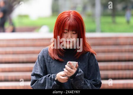 Jeune femme coréenne aux cheveux roux profondément absorbée par l'utilisation de son smartphone à l'extérieur Banque D'Images