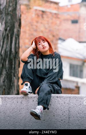Femme coréenne réfléchie avec des cheveux roux vibrants assis décontractés dans un lieu urbain Banque D'Images