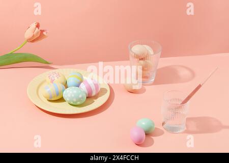 Oeufs de Pâques colorés décorés sur un plat jaune avec des tasses de fleurs et d'eau sur un fond rose pastel. Les œufs symbolisent la fertilité à Pâques. Main spéciale Banque D'Images