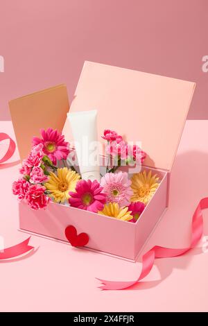 Un tube cosmétique blanc affiché sur une boîte-cadeau rose pleine de fleurs fraîches décorées avec une carte de papier brun et un ruban rose sur fond rose doux. Mocku Banque D'Images