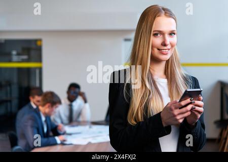 Femme d'affaires blonde confiante utilise un smartphone dans un bureau moderne, les collègues travaillent à table derrière. Une femme professionnelle en tenue décontractée élégante interagit avec Banque D'Images