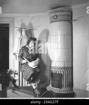 Dans les années 1940 Intérieur d'une pièce et une jeune femme avec un bol dans le genou qui fouette le contenu. Elle est assise à côté d'un poêle en faïence, une source de chauffage au bois qui était commune dans les maisons dans le passé. La chaleur a été stockée longtemps dans la grande masse du poêle. Suède 1946 Kristoffersson ref N138-3 Banque D'Images