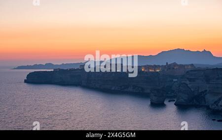 Bonifacio, Corse-du-Sud, Corse, France. Vue du sommet de la falaise à la citadelle illuminée, crépuscule, montagnes lointaines silhouettées sur un ciel doré. Banque D'Images