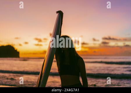 Jeune femme avec planche de surf debout à la plage contre le ciel pendant le coucher du soleil Banque D'Images