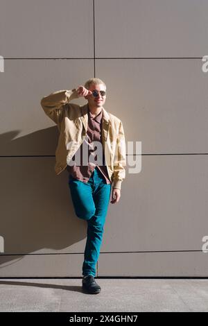 jeune homme gay avec des lunettes debout contre le mur au coucher du soleil, portant une veste marron et un pantalon en jean Banque D'Images