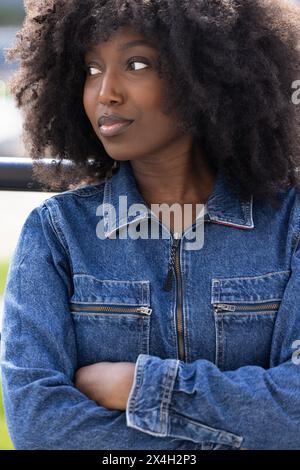 Un gros plan représente une femme noire avec un afro, vêtue d'une veste en denim classique, regardant avec une expression rêveuse. La mise au point douce sur son visage renforce son regard contemplatif, suggérant une pensée profonde ou une aspiration, dans un contexte urbain. Dreamy Black femme en veste denim regardant loin. Photo de haute qualité Banque D'Images